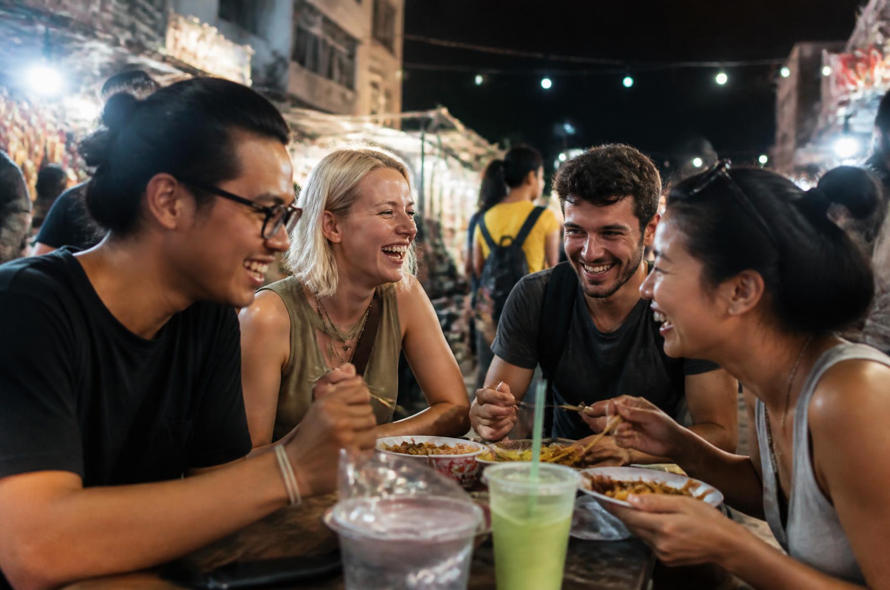 Viaggiatori Ferbi che ridono attorno a un tavolo di street food in un mercato notturno